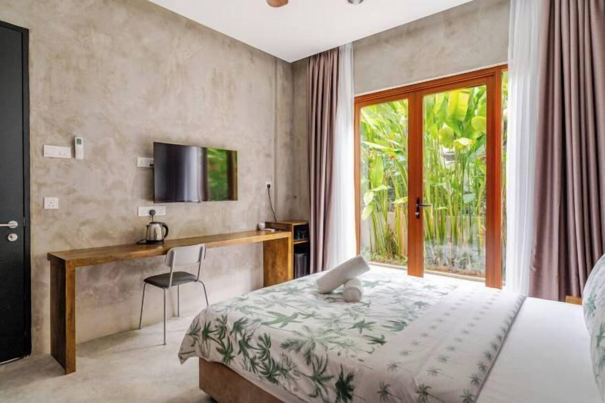 Un dormitorio con cama, escritorio, silla, televisor y una puerta corrediza de vidrio que da a un jardín.
