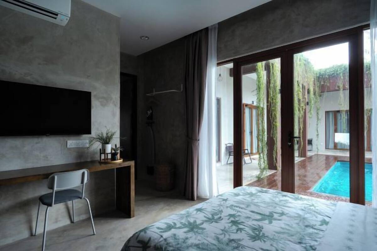 Un dormitorio moderno con una cama grande, un escritorio y un televisor. Tiene una puerta corrediza de vidrio que da a una zona de piscina.