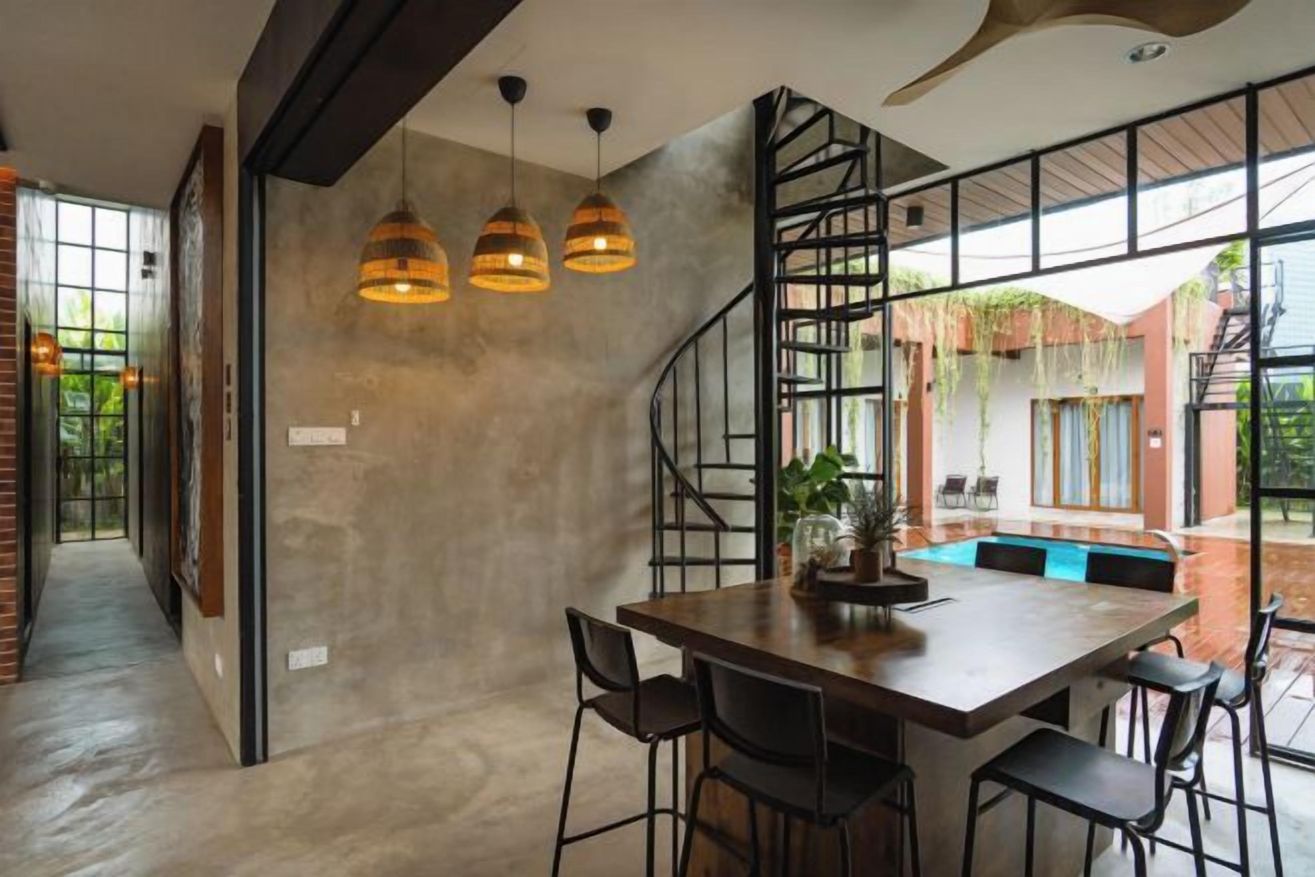 Un espace repas avec une table et des chaises en bois, des luminaires suspendus et une vue sur une piscine et un coin salon extérieur.