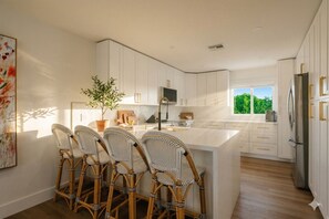 A modern kitchen with white cabinets, a central island, wicker chairs, and a large window. - Direct Oceanfront Luxury: Brand New Beach House at Coastal Therapy (St. Augustine)