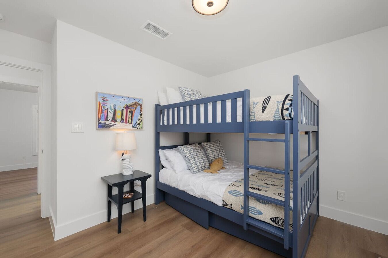 A bunk bed with a white and blue design, a small bedside table with a lamp, and a colorful painting on the wall.