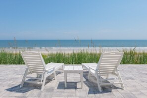Two white lounge chairs with a table on a paved patio overlooking a beach. - Direct Oceanfront Luxury: Brand New Beach House at Coastal Therapy (St. Augustine)