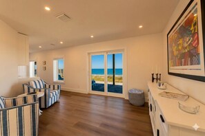 A spacious room with a large sliding glass door leading to a balcony with a view of the sea. There are striped chairs and a small round ottoman. - Direct Oceanfront Luxury: Brand New Beach House at Coastal Therapy (St. Augustine)
