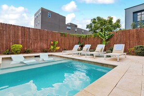 Ein rechteckiger Pool mit klarem, blauem Wasser, umgeben von einem Holzzaun und einigen Pflanzen. Vier Liegestühle mit Handtüchern stehen bereit, und im Hintergrund ist ein Gebäude mit einer grauen Fassade zu sehen.