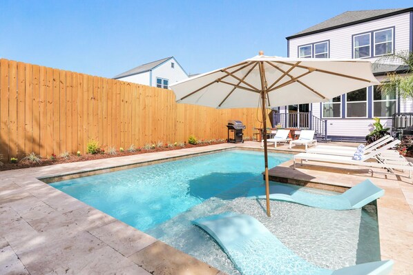 Ein Garten mit Schwimmbad, Holzzaun und einer Terrassenanlage mit Liegestühlen und Sonnenschirm.
