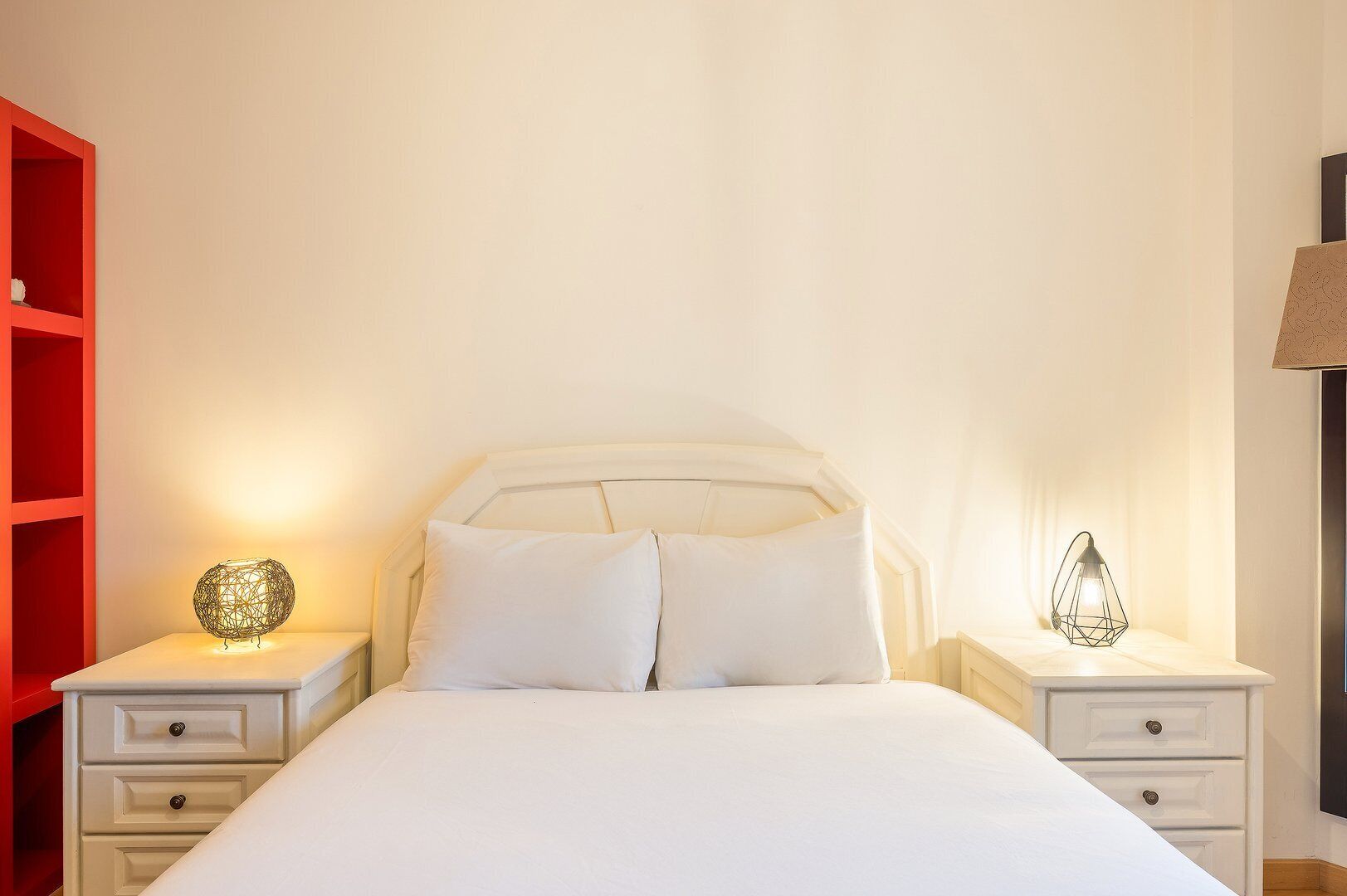 A neatly made bed with white linens, two bedside tables with lamps, and a red shelving unit.