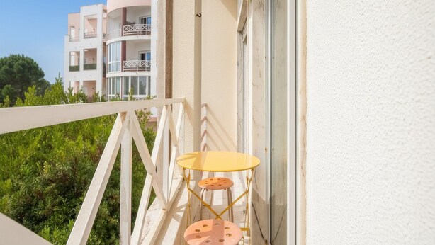 Ein Balkon mit weißer Brüstung, einem gelben Tisch und einem pinken Hocker.