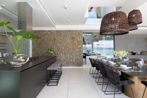 A modern kitchen with a stone wall, a dining area with chairs and a table, and a large island with a vase of flowers. - Syrah 3-Bedroom Beachfront Villa in Koh Samui (Koh Samui)