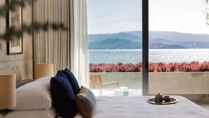 Une chambre avec un lit, des oreillers, une table de chevet avec une lampe, un plateau avec une théière, et une vue sur la mer et les montagnes.