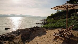 Une plage avec un transat en bois et un parasol, un rivage rocheux et une mer calme, avec des montagnes qui se dessinent à l’horizon.