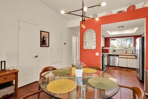 A dining area with a glass table, chairs with woven placemats, a pendant light fixture, and a kitchen in the background. - Sunset Vista Retreat (Palm Springs)