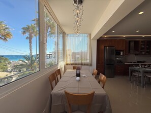 A dining area with a table set for two, large windows with a view of the beach, and a kitchen area with modern appliances. - Anna’s Seaside Apartment - In front of the beach (Kos)