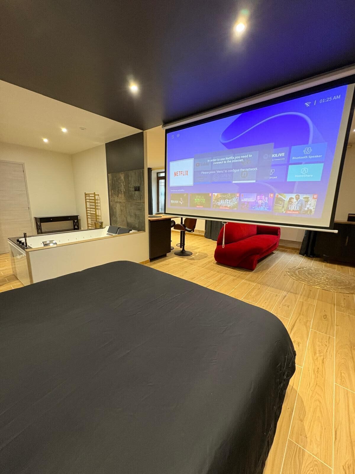 A modern hotel room with a large flat-screen TV displaying Netflix content, a bed with a dark cover, a red chair, and a wooden floor.