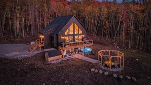 Une cabane dotée d’une grande terrasse, d’un bain à remous et d’un espace avec foyer, le tout entouré d’arbres.
