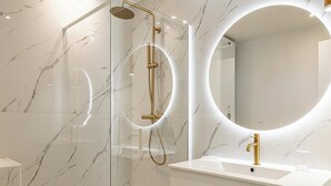 A modern bathroom with a glass-enclosed shower, a gold faucet, and a large mirror with a circular light. - Luxurious 2-bedroom with terrace 200m from Palais (Cannes)