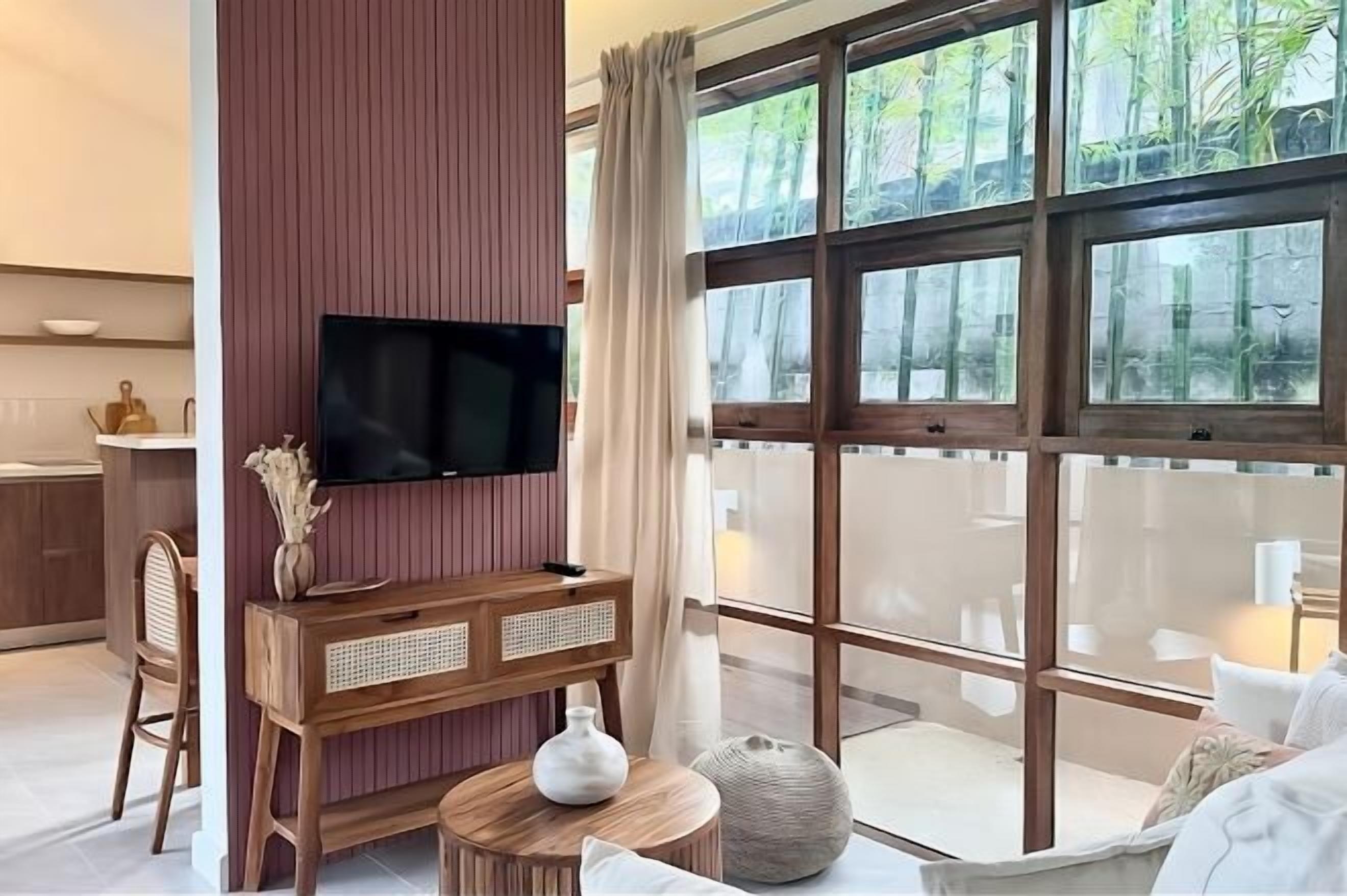 Una sala de estar moderna con un ventanal grande, un televisor de pantalla plana montado en un mueble de madera, y una zona de estar con una mesa redonda y cojines.