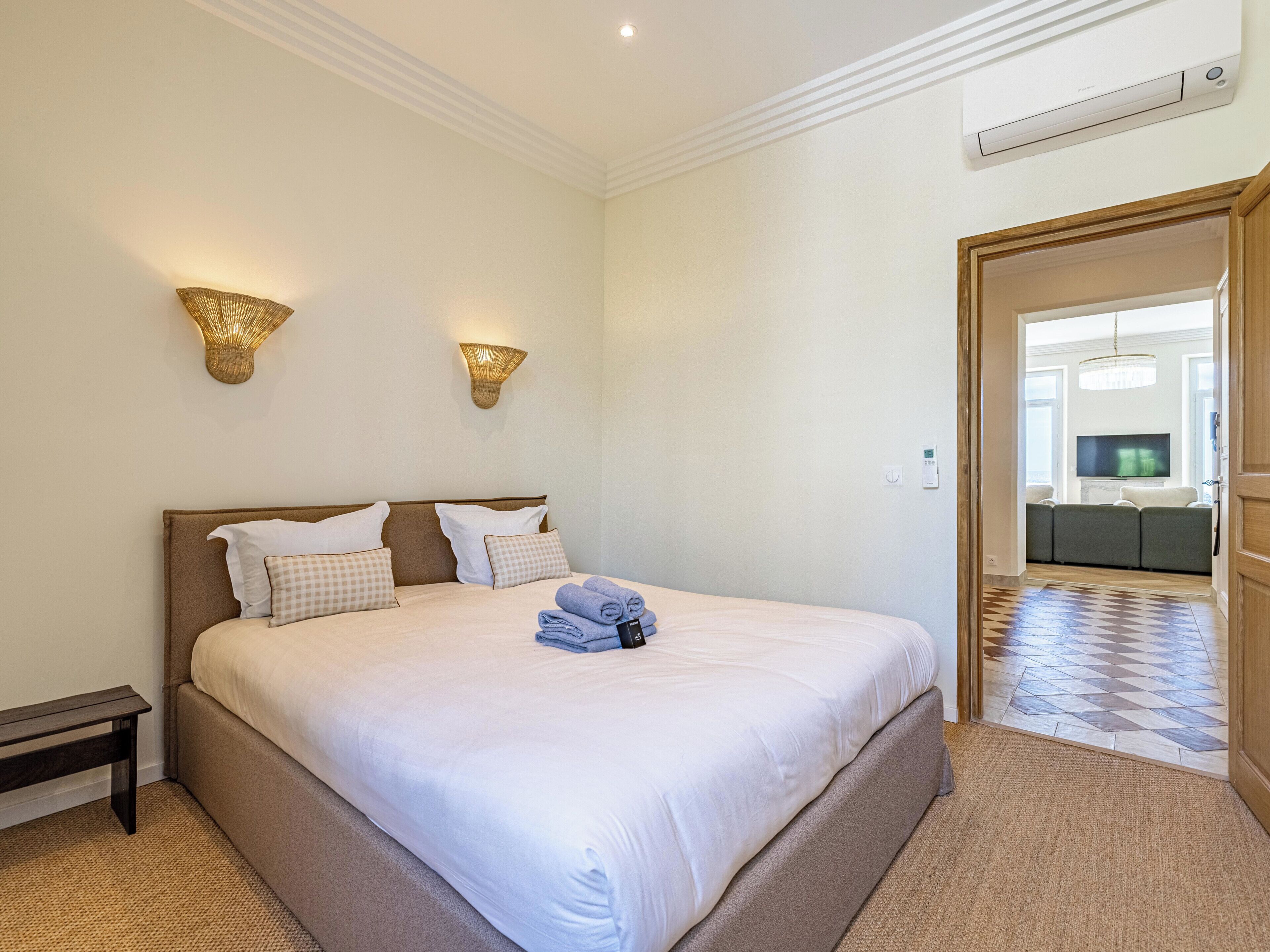 A hotel room with a large bed, two wall-mounted lamps, a flat-screen TV, and a view into the living area.
