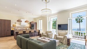 Ein modernes Wohnzimmer mit einer Couch, einem Esstisch und einem Fernseher. Es gibt eine Küchenzeile mit einer Bar und Blick aufs Meer.
