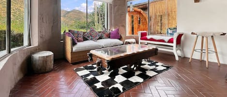 A room with a herringbone floor, a glass coffee table, a wicker sofa with colorful cushions, a red chair, and a view of mountains through large windows.