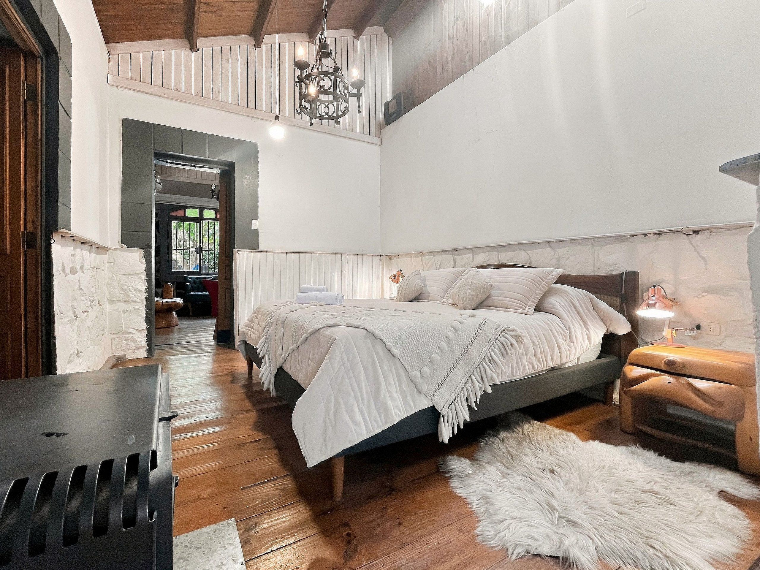 A bedroom with a large bed, wooden flooring, a chandelier, and a stone wall.