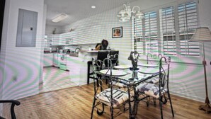 Un coin repas avec une table et des chaises, une cuisine à l’arrière-plan et une fenêtre avec des stores.