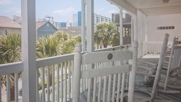 Une véranda couverte, dotée de balustrades blanches et de chaises en bois, qui donne sur un quartier résidentiel agrémenté de palmiers et de bâtiments.