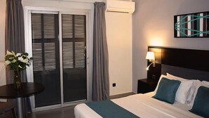 A hotel room with a bed, a nightstand, a lamp, a table with a vase of flowers, and a sliding door with curtains. - Hotel Farid (Dakar)