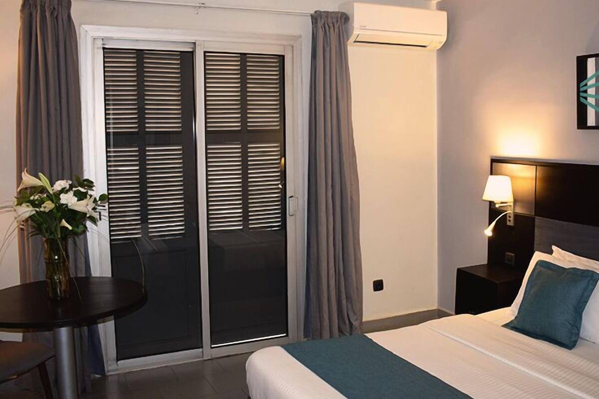 Una habitación de hotel con cama, mesita de noche, lámpara, una mesa con un jarrón de flores y una puerta corrediza con cortinas.