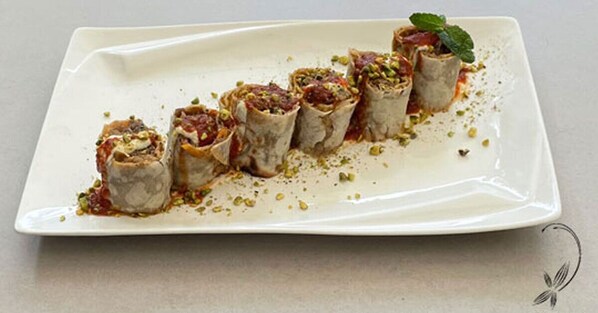 A white rectangular plate with six rolled pastries garnished with pistachios and a mint leaf, arranged in a row.