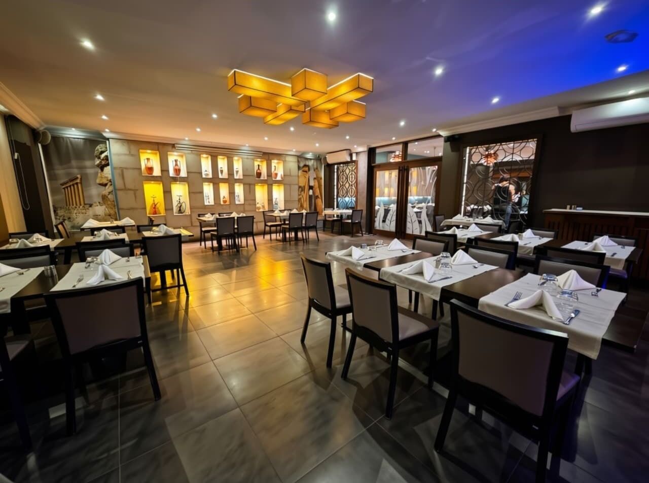 A restaurant with multiple tables set for dining, a modern ceiling design, and a wall with illuminated bottles.