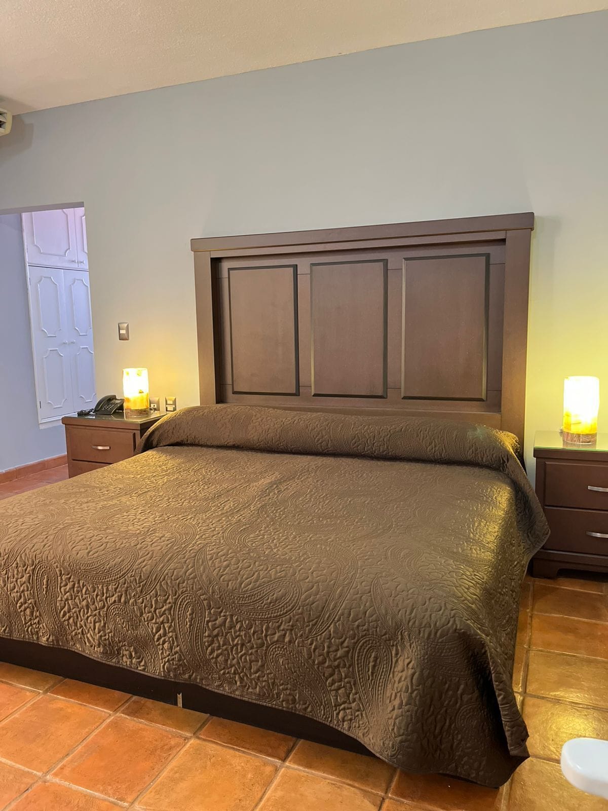 A hotel room with a large bed, a wooden headboard, two bedside tables with lamps, and a door with a decorative panel.