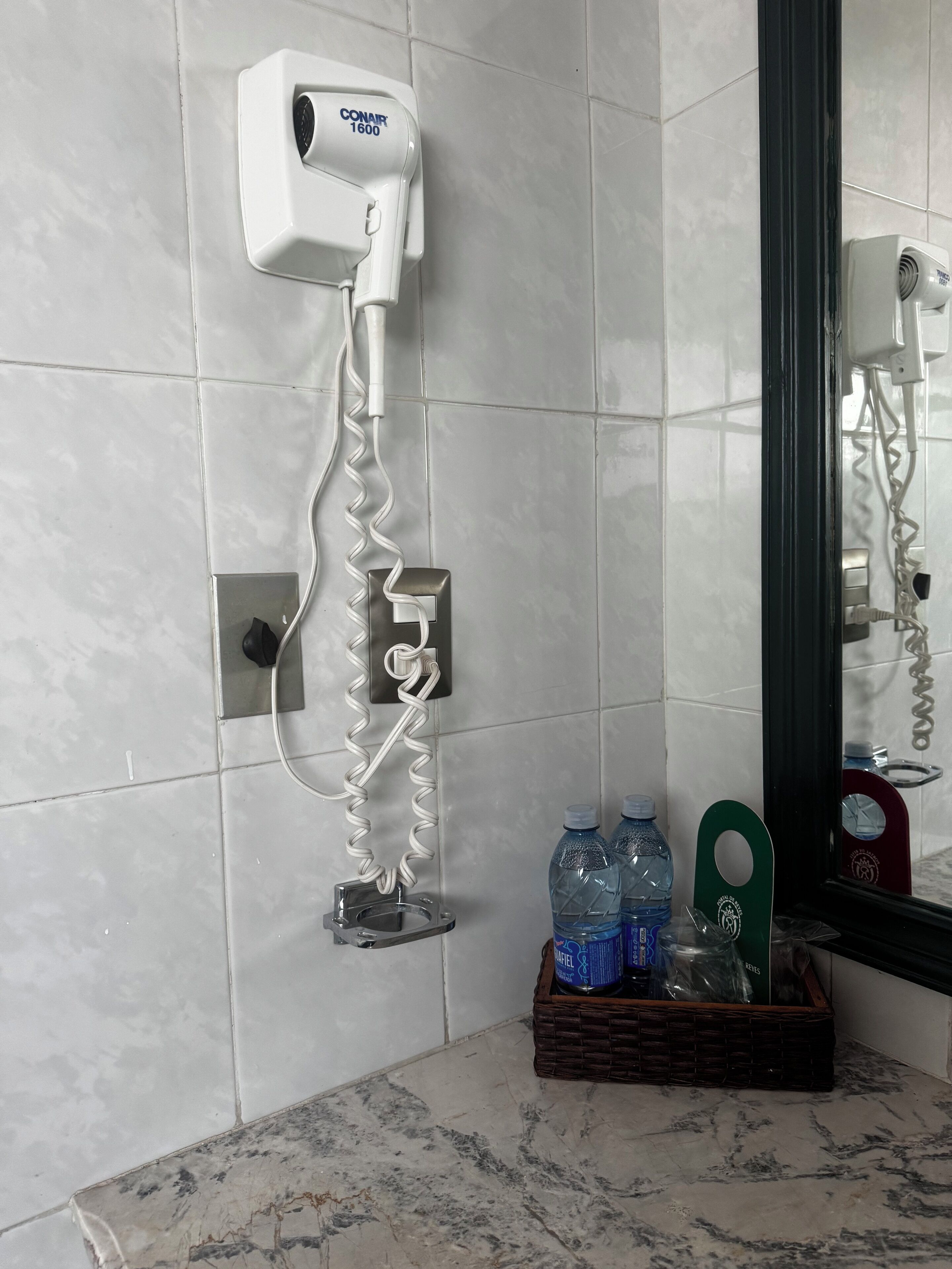 A bathroom with a hairdryer mounted on the wall, a mirror, and a basket containing bottled water and a green towel.