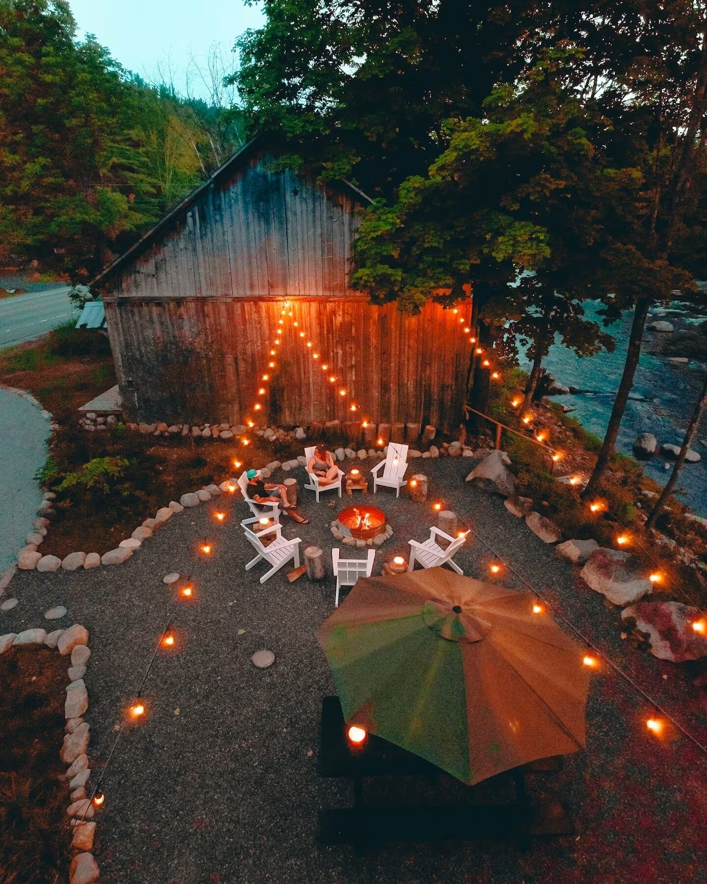 A rustic outdoor seating area with white chairs and a fire pit, surrounded by a stone border and string lights.