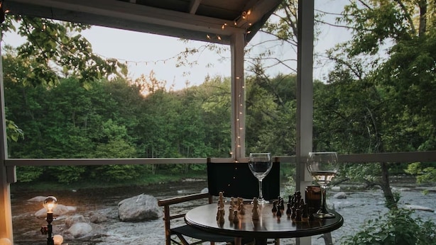 A wooden deck with a table set for two, overlooking a river. - Warner's Camp Riverfront Retreat: Cabins, Hot Tub, Saunas, Swimming Hole (Upper Jay)