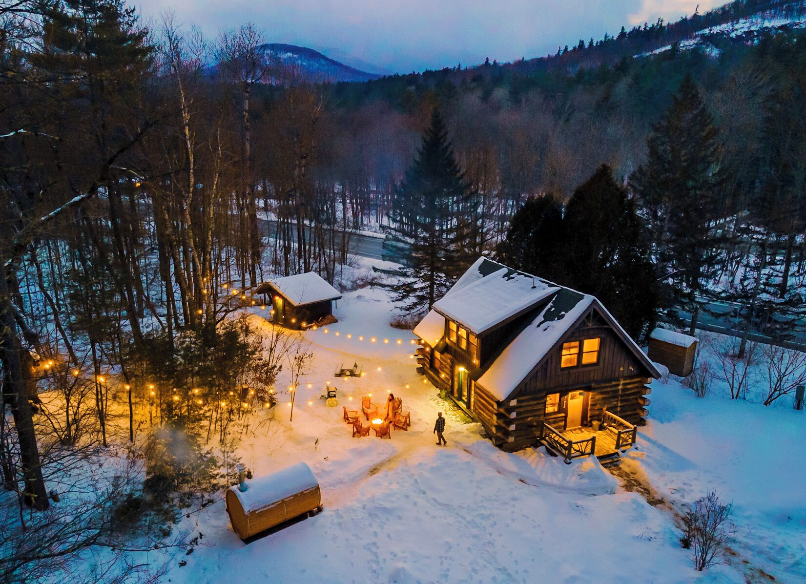 A log cabin with a snow-covered roof, surrounded by a forest and a mountain in the background.