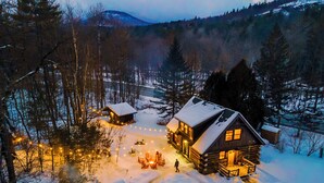 A log cabin with a snow-covered roof, surrounded by a forest and a mountain in the background. - Warner's Camp Riverfront Retreat: Cabins, Hot Tub, Saunas, Swimming Hole (Upper Jay)