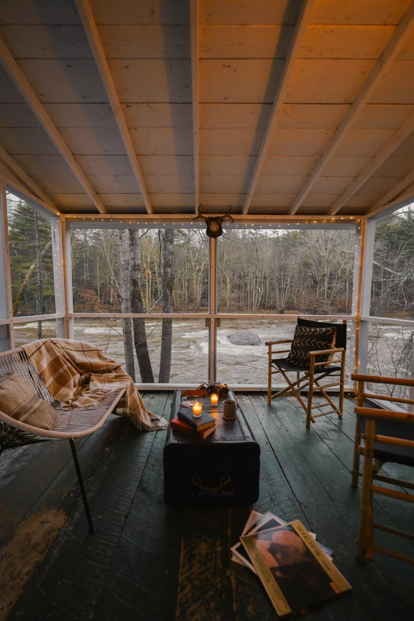 Une pièce chaleureuse avec un plafond en bois, une grande fenêtre donnant sur une forêt, deux fauteuils en osier, une petite table avec une bougie et une boîte sur laquelle est écrit : « J’AI BATTU LE NA ».