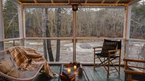 A cozy room with a wooden ceiling, a large window overlooking a forest, two wicker chairs, a small table with a candle, and a box labeled "I BEAT THE NA". - Warner's Camp Riverfront Retreat: Cabins, Hot Tub, Saunas, Swimming Hole (Upper Jay)