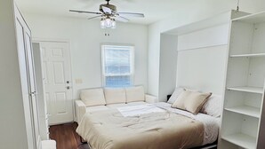A bedroom with a bed, a ceiling fan, a window with blinds, a door, and a built-in shelving unit. - The Starland Loft Apartment- Savannah, GA (Savannah)