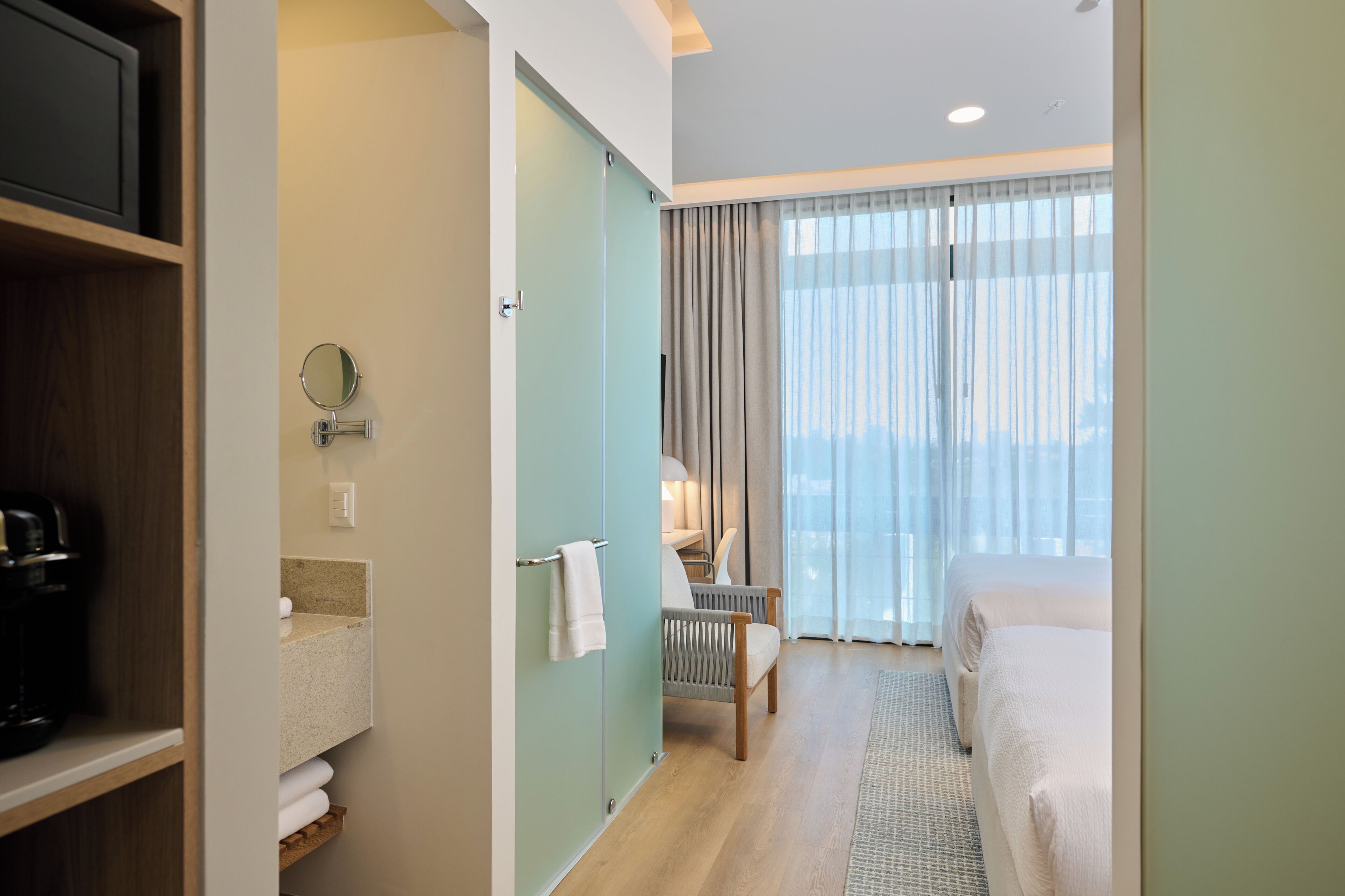 A hotel room with a bed, a chair, a desk with a lamp, a mirror, and a view of the city through large windows.