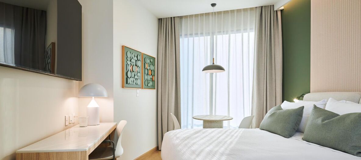 A hotel room with a bed, desk, chair, lamp, and artwork on the wall.