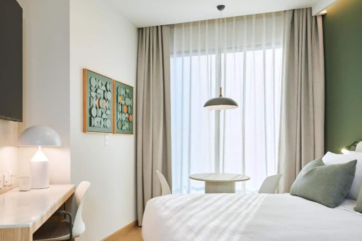 Quarto de hotel com cama, escrivaninha, cadeira, abajur e obras de arte na parede.