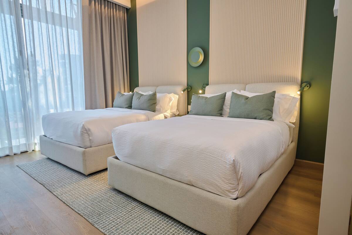 Quarto de casal com duas camas, uma janela grande com cortinas e uma parede verde decorativa.