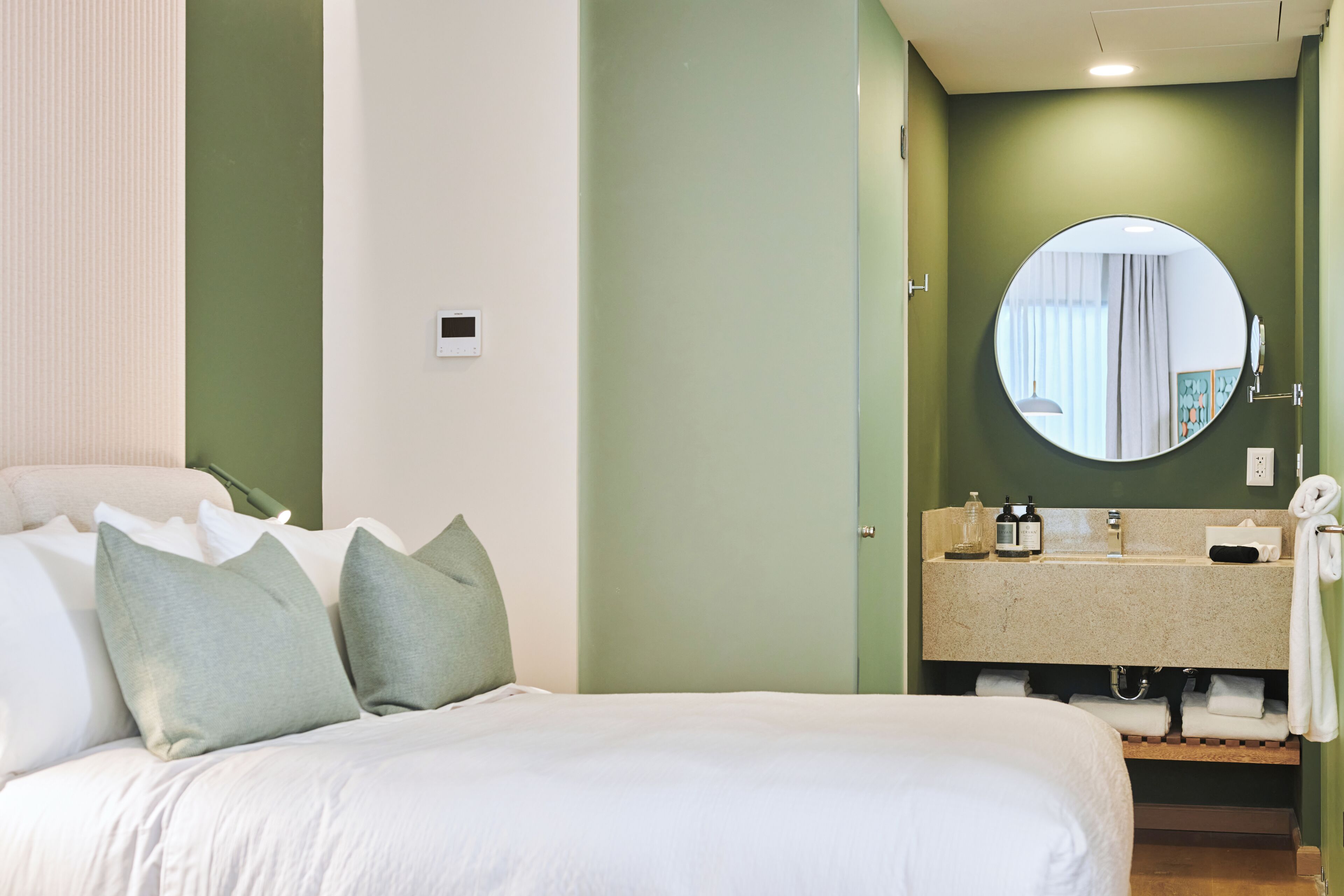 Une chambre d’hôtel avec un lit, des oreillers, un miroir rond, un lavabo et un porte-serviettes.