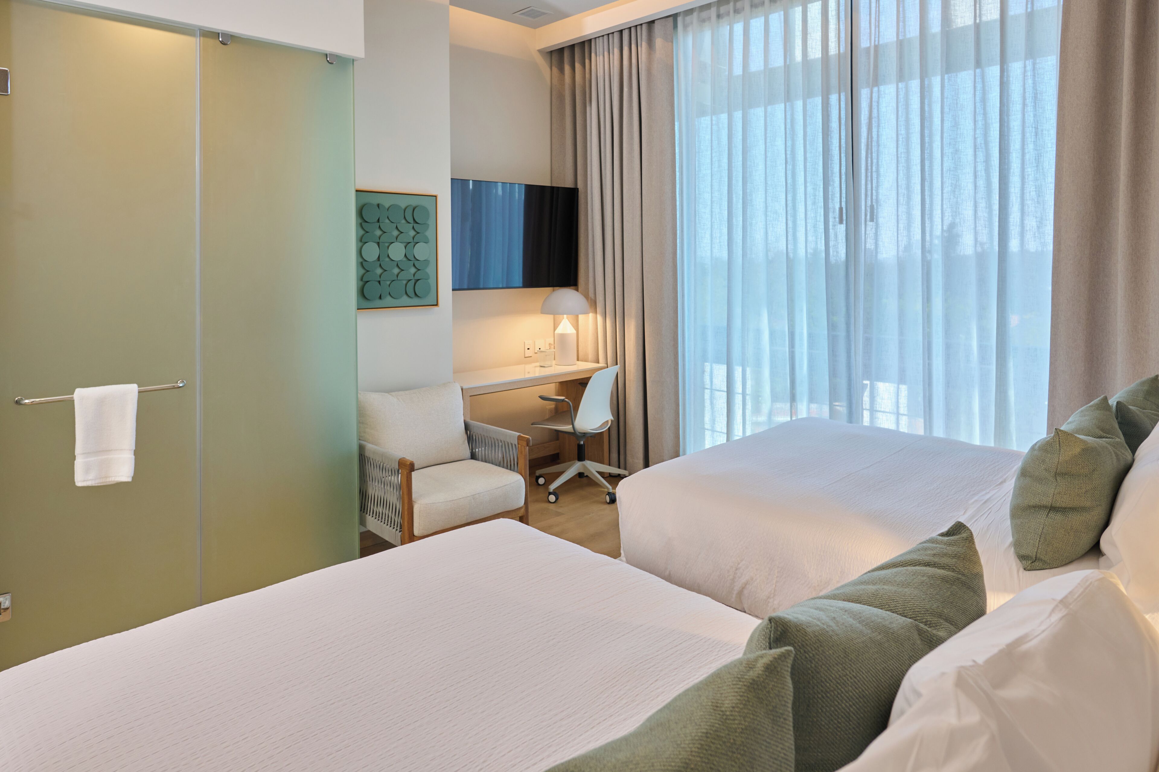 Une chambre d’hôtel avec deux lits, un bureau, une chaise, une lampe et une grande fenêtre avec des rideaux.