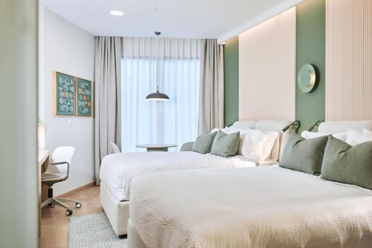 Ein Hotelzimmer mit zwei Betten, einem Schreibtisch, einem Stuhl, einem fenster mit Vorhängen und einer Wanduhr.