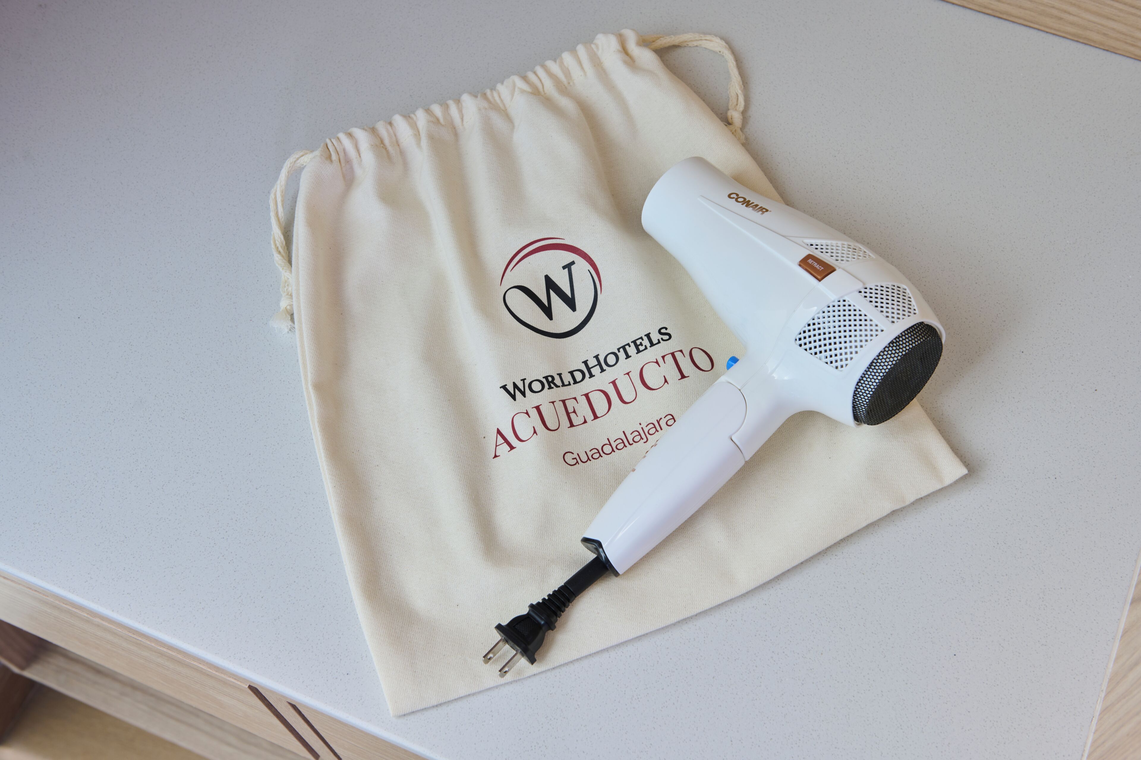 A white hair dryer with a cord plugged into it, placed on a beige drawstring bag with the text 'WorldHotels Acueducto Guadalajara' visible.