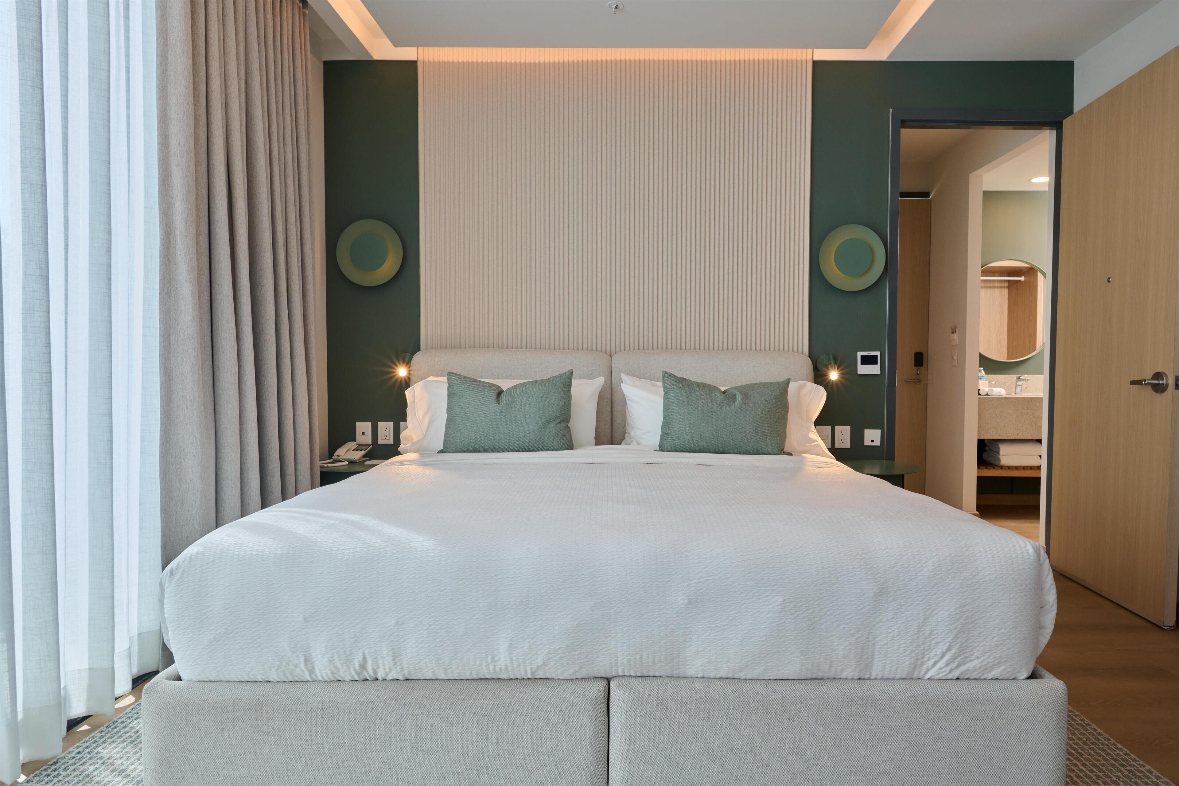 Een net opgemaakt bed met een wit en grijs kleurenschema, een nachtkastje met een lamp en een zicht op een badkamer.