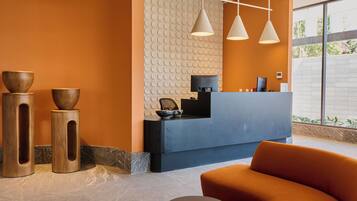 Un área de recepción con un mostrador azul, tres lámparas colgantes y una zona de estar con un sofá naranja y una mesa redonda.
