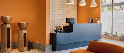 Een receptiegedeelte met een blauwe balie, drie hanglampen en een zithoek met een oranje bank en een ronde tafel.